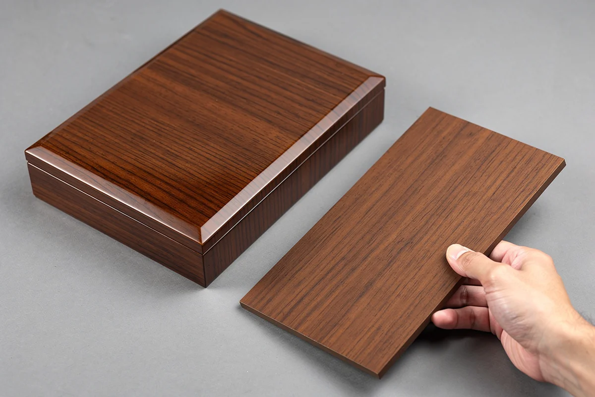 A hand holding a wood-grain sample panel beside a polished wooden box